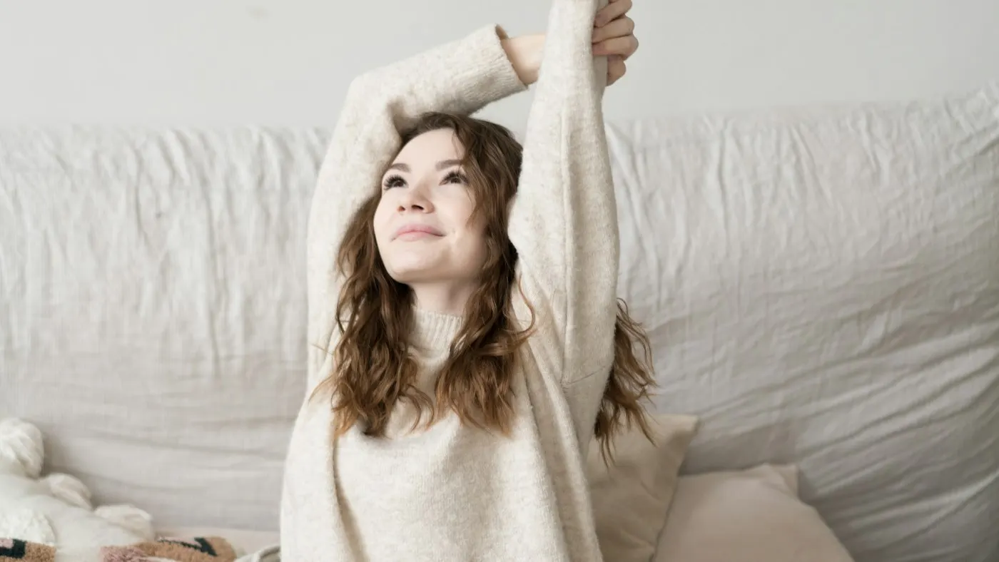 A woman in a cream sweater sitting up in bed and stretching both arms overhead in soft morning light