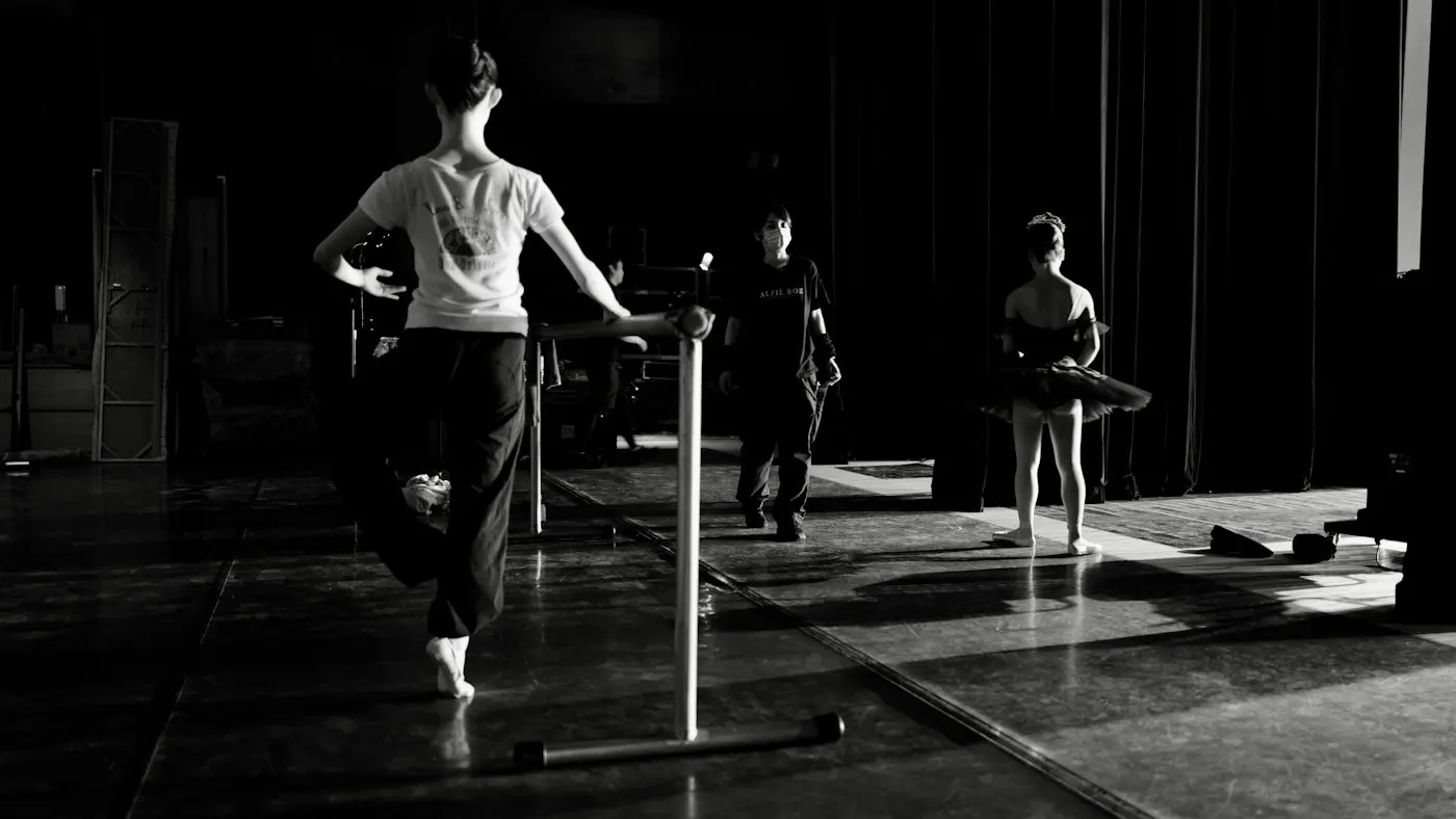 Ballet dancers practicing at a barre in a studio, one balancing on the ball of her foot in black and white
