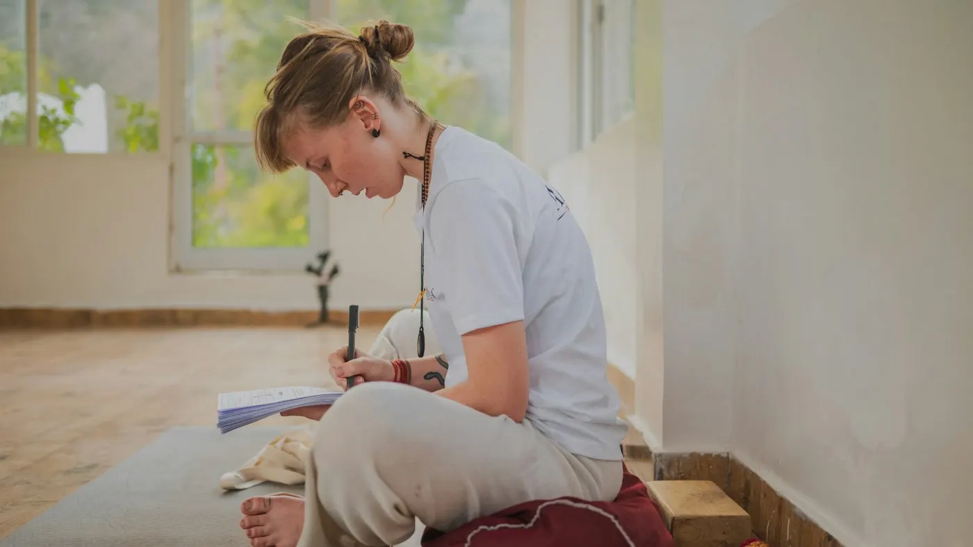 A woman sits cross-legged on a cushion beside an exercise mat in a bright practice studio, taking notes in an open notebook