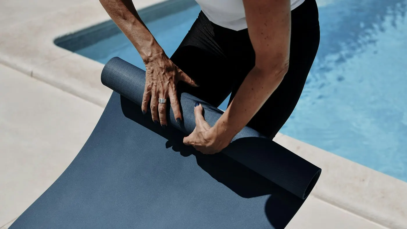 Hands rolling up a thin dark-blue exercise mat on a sunny pool deck