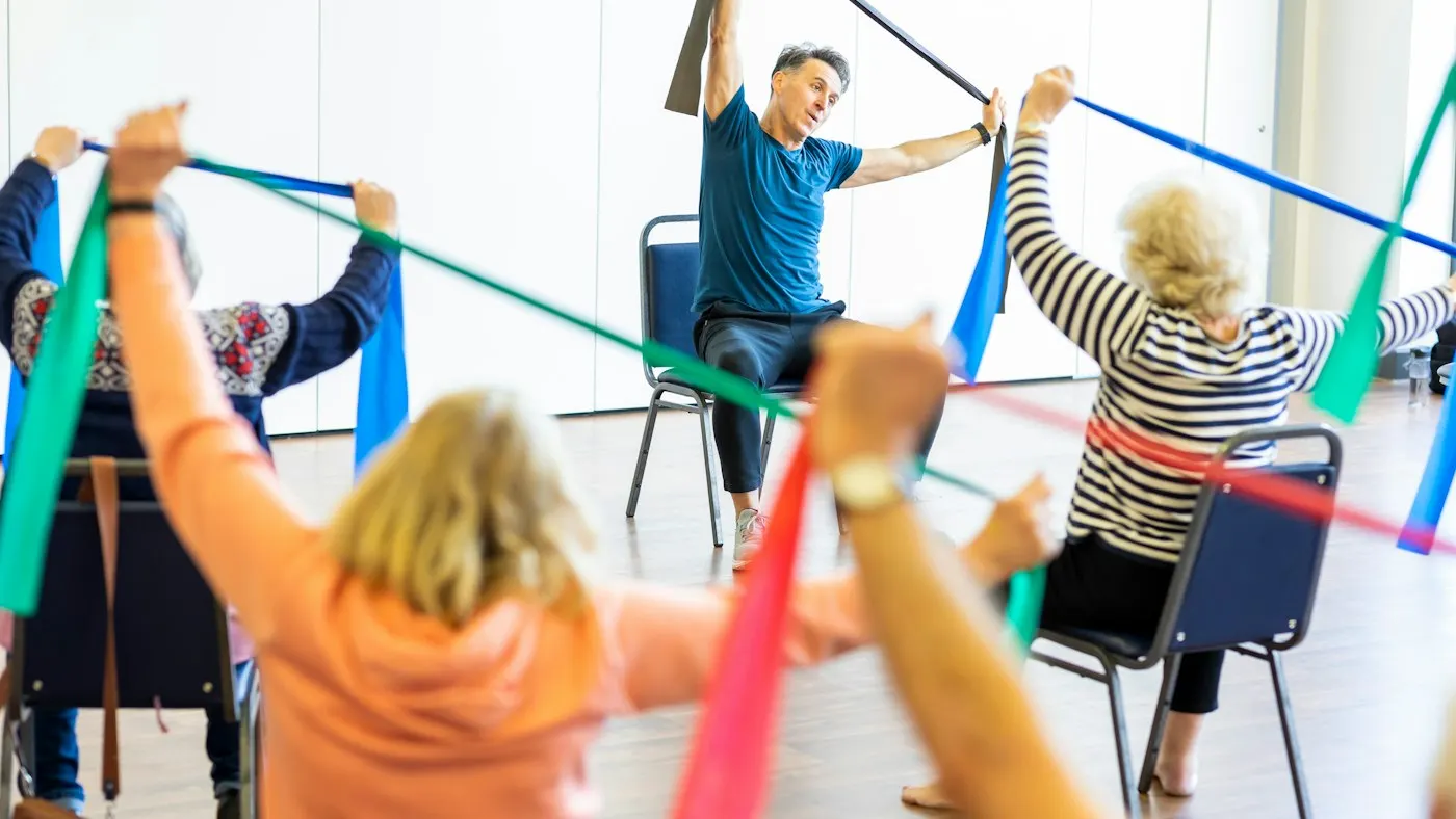 Older adults sitting on armless chairs in a community exercise class, stretching resistance bands overhead while an instructor leads from a chair