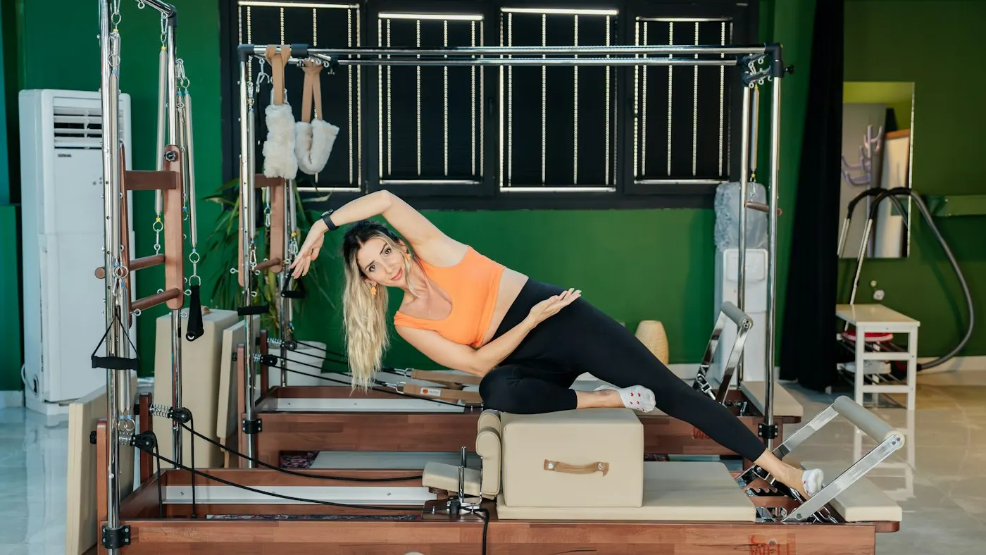 A woman stretches on a Pilates reformer in a studio with wooden reformers along a green wall