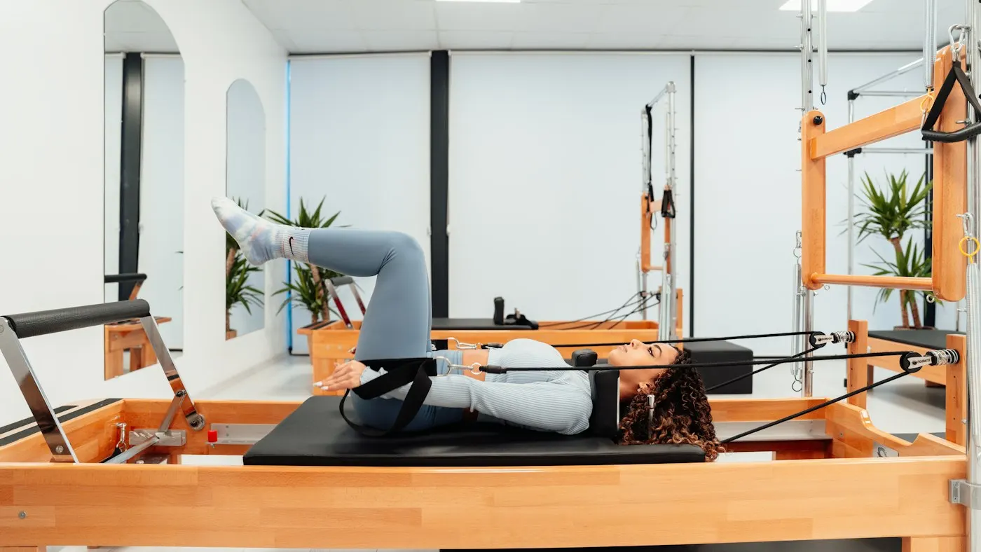 Woman in a fitted top, leggings and grip socks doing a legs-up exercise on a wooden Pilates reformer in a studio